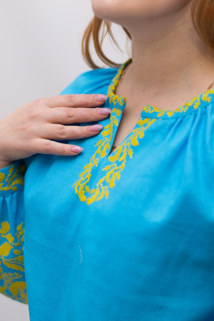 Close-up of a woman wearing a blue blouse with traditional Ukrainian embroidery, hand on chest.
