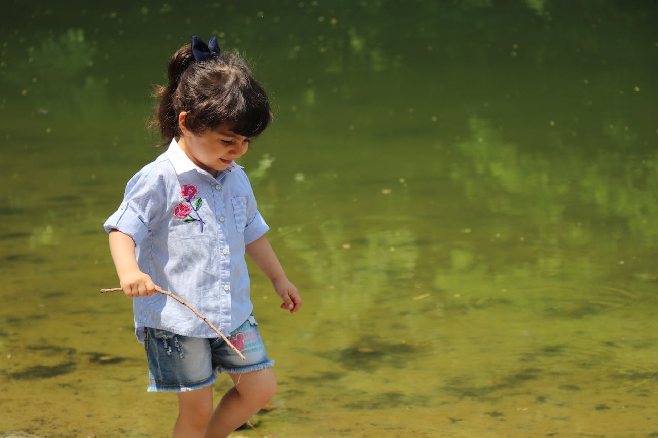 A young girl playing by a lake with a stick, enjoying summer outdoors.